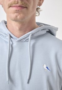 Light blue hoodie in soft fabric featuring a front kangaroo pocket, drawstring hood, and a small embroidered bird detail in white and blue.