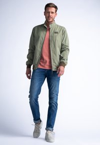 Světlá olivově zelená bomber bunda s stojáčkem, hnědými akcenty a kapsami na zip, nošená přes korálovou košili a modré džíny.