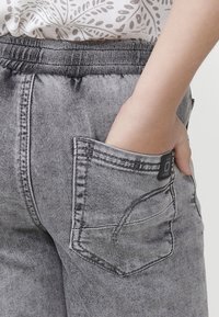 Pantalones de mezclilla gris con una cintura elástica y un bolsillo lateral, con costuras de contraste y una pequeña etiqueta de marca en el bolsillo.