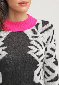 Nainen, jolla on päällään mustavalkoinen kuvioitu neulepaita, jossa on kirkkaan vaaleanpunainen ribattu pyöreä pääntie, näkyvillä hartioista huuliin.