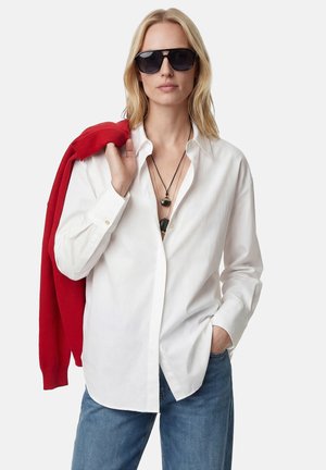 Blondynka w czarnych okularach przeciwsłonecznych, białej koszuli zapinanej na guziki, z warstwowymi naszyjnikami, w niebieskich dżinsach, trzymająca czerwony sweter na ramieniu.