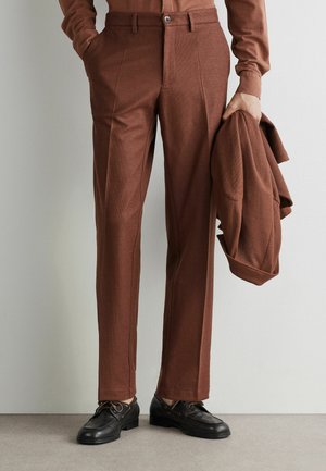 Mann trägt braune maßgeschneiderte Hose und passendes Hemd, hält eine braune Jacke in der rechten Hand und trägt schwarze Lederschuhe.