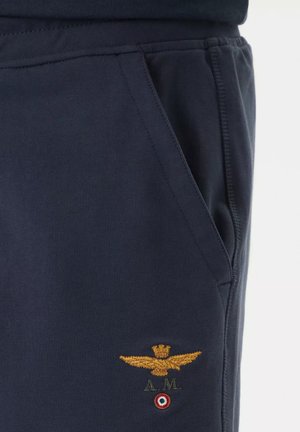 Tasca dei pantaloni blu scuro con aquila dorata ricamata, corona, iniziali "A.M." ed emblema circolare rosso, bianco e verde sotto il ricamo.