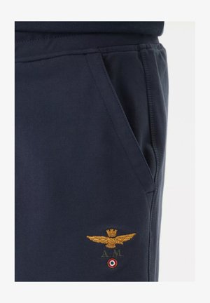 Tasca dei pantaloni blu scuro con aquila dorata ricamata, corona, iniziali "A.M." ed emblema circolare rosso, bianco e verde sotto il ricamo.