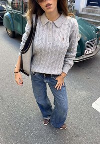 Žena v sivom svetríku s vrkočovým vzorom, široké nohavice, hnedé topánky, nesie tašku, stojí na ulici pri vintage zelenom aute.