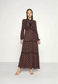 Mulher vestindo um longo vestido preto floral com mangas longas e sandálias de salto alto bege, de pé contra um fundo branco simples.