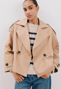 Beige oversized trenchcoat met dubbelgeknoopte knopen, brede revers en schouderbanden, gelaagd over een crème en marineblauwe gestreepte trui.