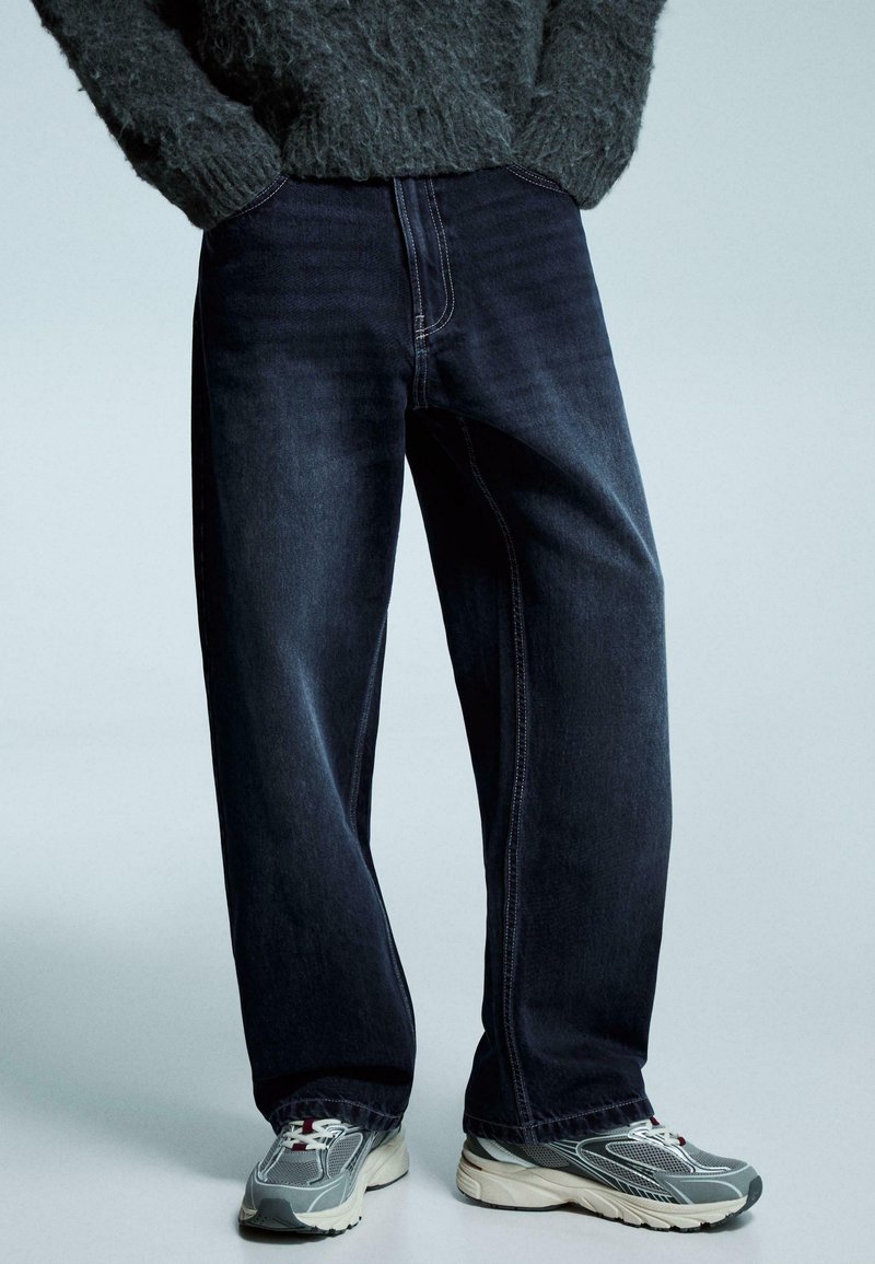 Dunkelblaue Jeans mit geradem Bein und kontrastierender Naht, kombiniert mit grauen und cremefarbenen Sportschuhen. Die Textur wirkt glatt und strapazierfähig.