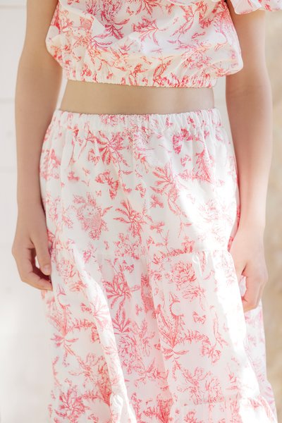 Conjunto de blusa e saia de tecido branco com estampa floral vermelha, apresentando uma cintura elástica e bainha com babados, textura suave e corte descontraído.