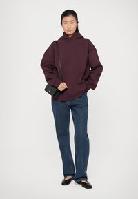 Felpa oversize bordeaux in tessuto morbido con tasca frontale, abbinata a jeans blu a gamba dritta e ballerine nere, mentre tiene una piccola borsa nera.