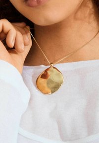 Close-up van een persoon die een witte top draagt, met een gouden, onregelmatig gevormd hangertje aan een delicate ketting bij hun hals.