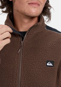 Chaqueta de forro polar marrón con cierre delantero, superficie texturizada y hombros en contraste en color marino. Presenta un pequeño parche con logo en el pecho.
