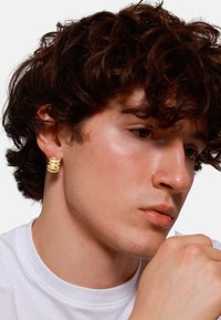 Orecchino a cerchio dorato a spirale su un modello. L'orecchino presenta una superficie liscia e lucida e un design arrotondato. Il modello indossa una camicia bianca.