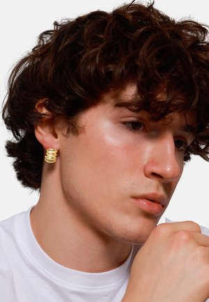 Boucle d'oreille en spirale dorée sur un mannequin. La boucle présente une surface lisse et brillante ainsi qu'un design arrondi. Le mannequin porte une chemise blanche.