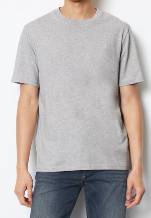 Mężczyzna w prostym, jasnoszarym t-shircie z krótkim rękawem i niebieskich dżinsach, stojący na białym tle.