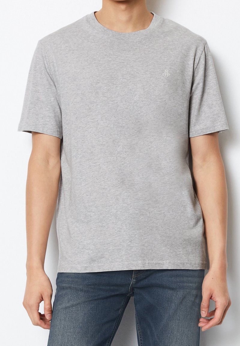 Homme portant un t-shirt gris clair uni à manches courtes et un jean bleu, debout devant un fond blanc.