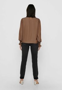 Brązowa koszula z długim rękawem i bufiastymi ramionami, z detalem w kształcie klucza na plecach oraz teksturowanymi mankietami, w zestawieniu z czarnymi dżinsami i przezroczystymi sandałami na obcasie.