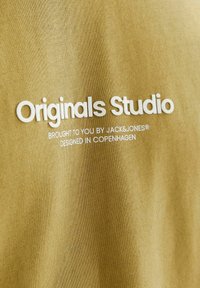Olive green fabric featuring textured knit, with white lettering reading "Originals Studio" and smaller text below in a simple font.
