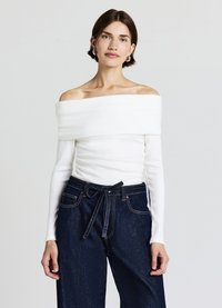 Weißes Schulterfreies, geripptes Oberteil mit langen Ärmeln, kombiniert mit hoch taillierter dunkler Jeanshose mit gebundener Gürtelschnalle und sichtbaren Nähten.