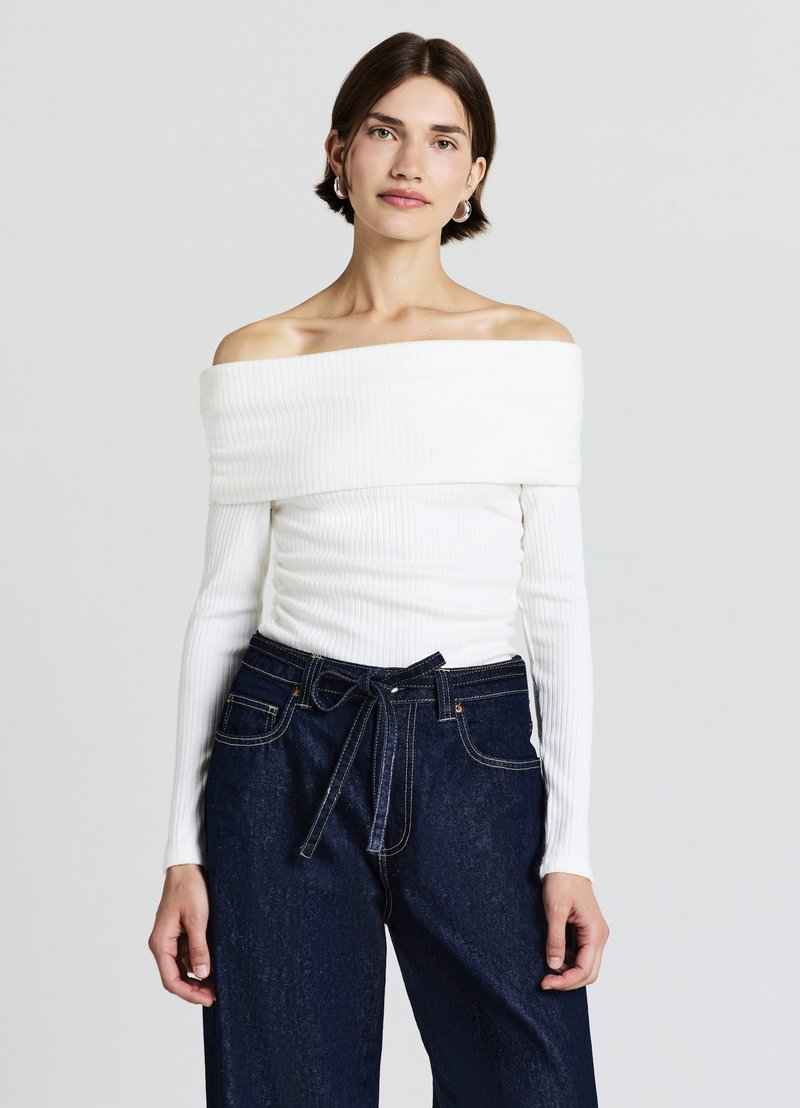 Weißes Schulterfreies, geripptes Oberteil mit langen Ärmeln, kombiniert mit hoch taillierter dunkler Jeanshose mit gebundener Gürtelschnalle und sichtbaren Nähten.