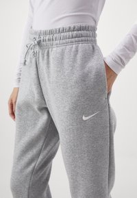 Osoba w jasnoszarych spodniach dresowych Nike z wiązaniem w pasie i białym logo Nike, z ręką w kieszeni, w połączeniu z białą bluzą z długim rękawem.