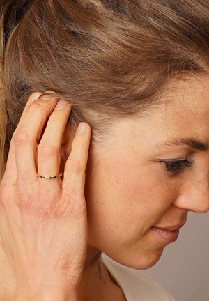 Goldring mit einem strukturierten, gewellten Design, getragen am dünnen Finger. Die Hand ist nah am Ohr platziert, während lange Haare herunterfallen.