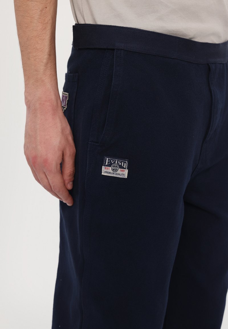 Pantalones de chándal de algodón azul marino con bolsillos laterales y cinturilla tejida. Parche de la marca Evisu notable con la etiqueta "EST. 1991" en la pierna.