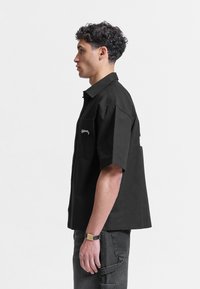 Jeune homme debout de profil, portant une chemise noire ample à manches courtes et un pantalon foncé avec une montre au poignet, sur un fond uni.