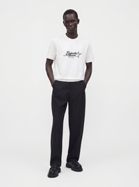 Άνδρας με λευκό T-shirt Emporio Armani, μαύρο παντελόνι και μαύρα παπούτσια στέκεται με τα χέρια στις τσέπες σε λευκό φόντο.