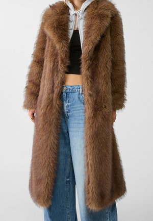 Person iført en lang, brun pelsjakke over en grå hættetrøje og sort crop top, kombineret med højtaljede blå jeans foran en ensfarvet baggrund.