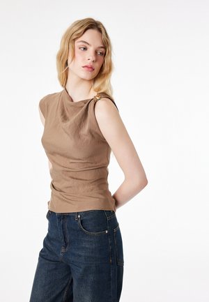 Młoda kobieta o blond włosach, ubrana w brązowy top bez rękawów z ozdobą w postaci złotej klamry oraz ciemnoniebieskie dżinsy, patrząca w bok.