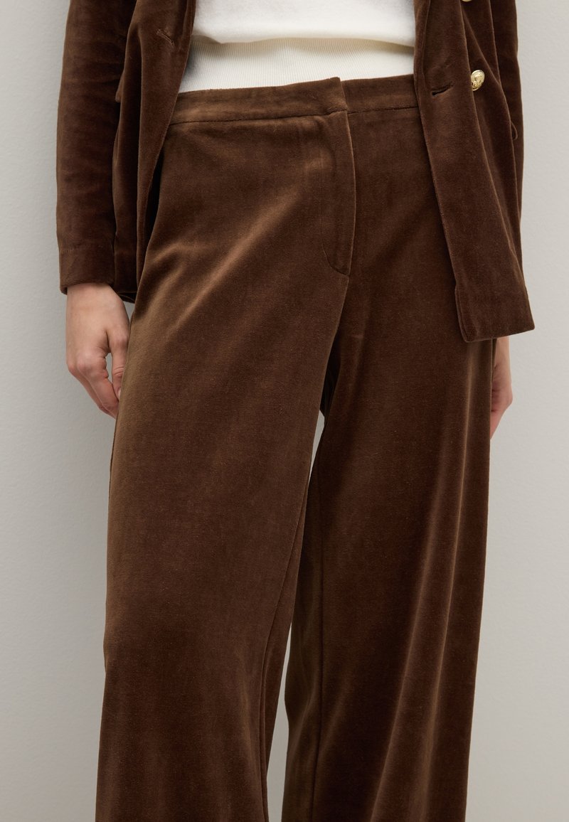 Pantaloni larghi in velluto marrone con una texture liscia, dotati di tasche frontali e una vestibilità rilassata, abbinati a un blazer coordinato.