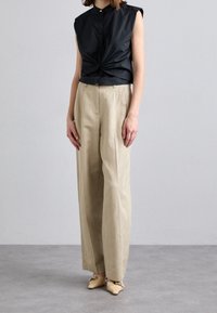 Camisa negra sin mangas con botón en la parte delantera y un diseño torcido, combinada con pantalones de pierna ancha beige y zapatos de color claro. Textura de tela suave visible.