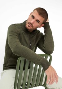 Dunkelgrüner Strickpullover mit Reißverschluss vorne, gerippten Bündchen und Seitendetails. Getragen mit hellfarbigen, strukturierten Hosen, sitzend auf einem grünen Stuhl.