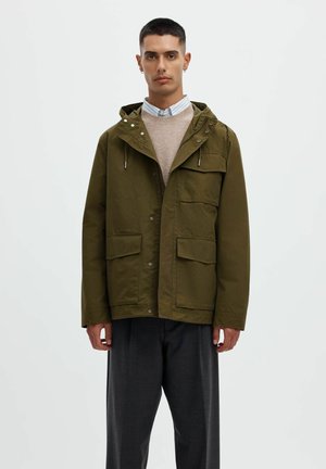 Jeune homme portant une veste à capuche vert olive avec poches à rabat, un pull beige, une chemise rayée à col et un pantalon gris foncé, debout devant un fond blanc.