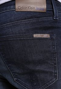 Tmavě modré džínové kalhoty s jemnou texturou. Obsahují hnědý kožený štítek na pásu a malou značkovou cedulku na zadním kapse.