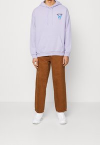 Jasnofioletowa bluza z kapturem, z kieszenią z przodu i graficznym wzorem, zestawiona z brązowymi spodniami o szerokich nogawkach oraz białymi trampkami.