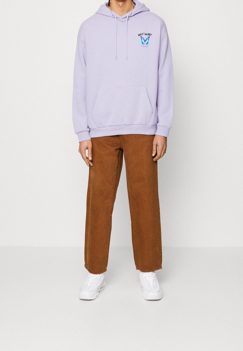 Jasnofioletowa bluza z kapturem, z kieszenią z przodu i graficznym wzorem, zestawiona z brązowymi spodniami o szerokich nogawkach oraz białymi trampkami.