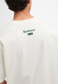 Kreemivärvi T-särk, mis on valmistatud puuvillast. Seljaosal on roheline "Barbour" logo ja graafika. Mugav lõige ning ümmargune kaelus.