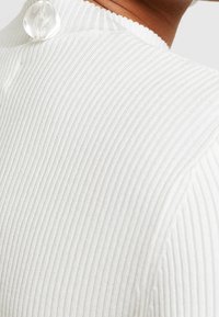 Weißes, geripptes Oberteil mit hohem Kragen und transparentem Knopfdetail am Rücken. Die Textur ist glatt mit dezenten vertikalen Linien.