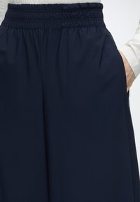 Personne portant un pantalon bleu marine avec une taille élastique, main droite dans la poche, chemise blanche à manches longues visible.