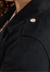 Veste en daim noir avec un col structuré, de grandes épaulettes hexagonales et un détail de bouton pression argenté.