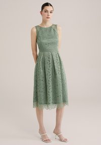 Grön spetsklänning med en åtsittande bodice, A-linjeformad kjol och scalloped nederkant. Har intrikata mönster, ärmlös design och en definierad midja.