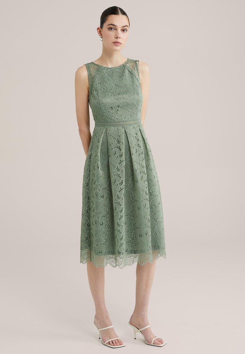 Grön spetsklänning med en åtsittande bodice, A-linjeformad kjol och scalloped nederkant. Har intrikata mönster, ärmlös design och en definierad midja.