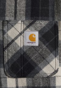 Grau und schwarze karierte Flanellstoff mit strukturierter Oberfläche. Verfügt über ein weißes Etikett mit einem gelben Logo und einer quadratischen Tasche.