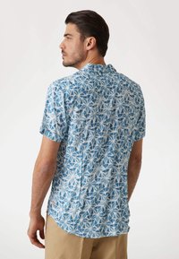 Camisa de manga corta en azul claro con un patrón geométrico blanco. Cuenta con un cuello de punta, un corte relajado y un dobladillo curvado.