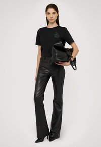 Kobieta w czarnych skórzanych spodniach i czarnym t-shircie, trzymająca dużą czarną skórzaną torbę na tle jednolitym.
