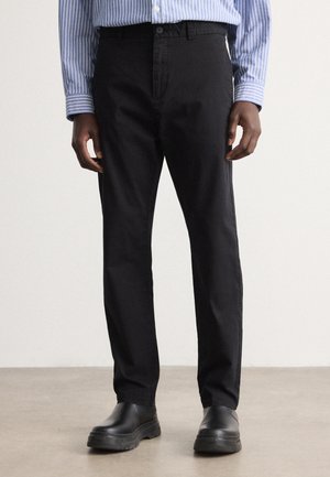 Mężczyzna ubrany w czarne spodnie o prostych nogawkach, czarne wsuwane buty oraz niebiesko-białą koszulę w paski zapinaną na guziki, stojący na szarej podłodze.