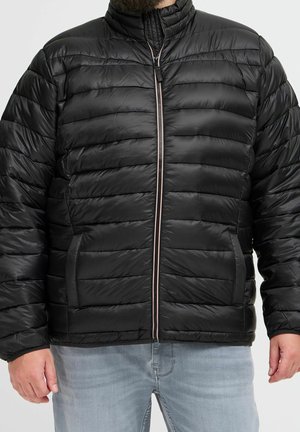 Man wearing a black quilted puffer jacket with front zipper and side pockets, paired with light blue jeans, standing against plain background.