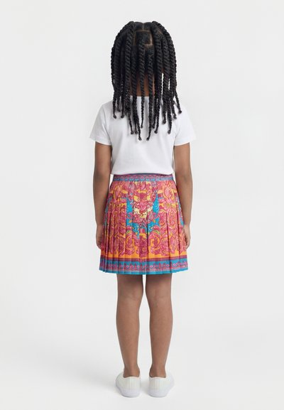 Παιδί με στριμμένα κοτσιδάκια, που φοράει άσπρο μπλουζάκι, πολύχρωμη φούστα με πτυχώσεις και άσπρα αθλητικά παπούτσια, στέκεται γυρισμένο πλάτη σε λευκό φόντο.